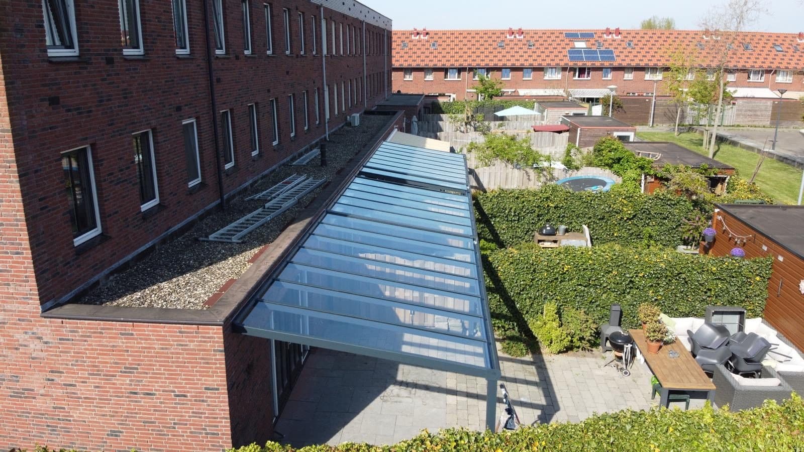 Long toit en verre anthracite adossé à une maison de ville en brique, vu d'en haut, au-dessus d'une terrasse et d'un salon de jardin