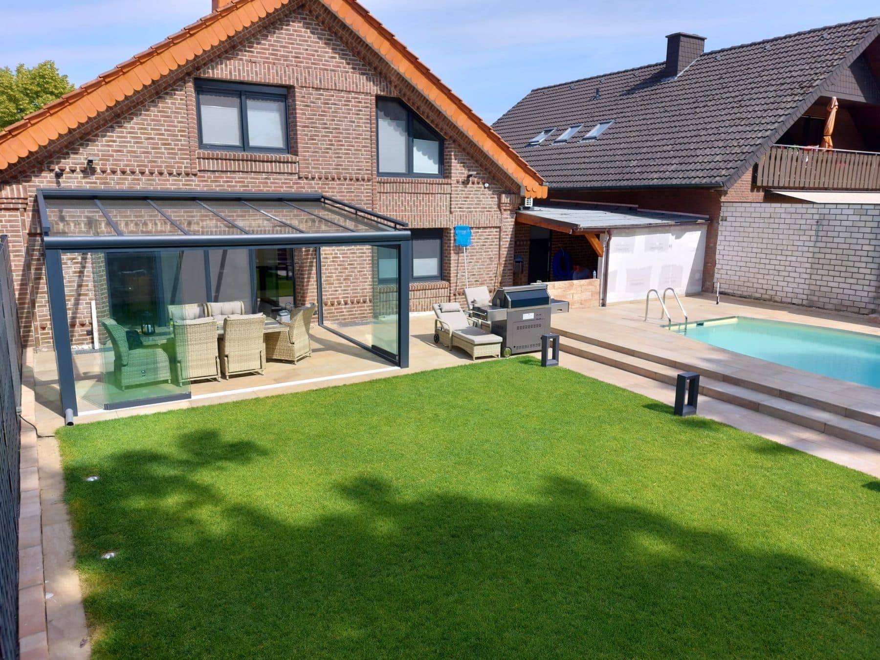 Pergola anthracite à toit en verre adossée à une maison en brique, avec salon de jardin, à côté d'une piscine et d'une pelouse