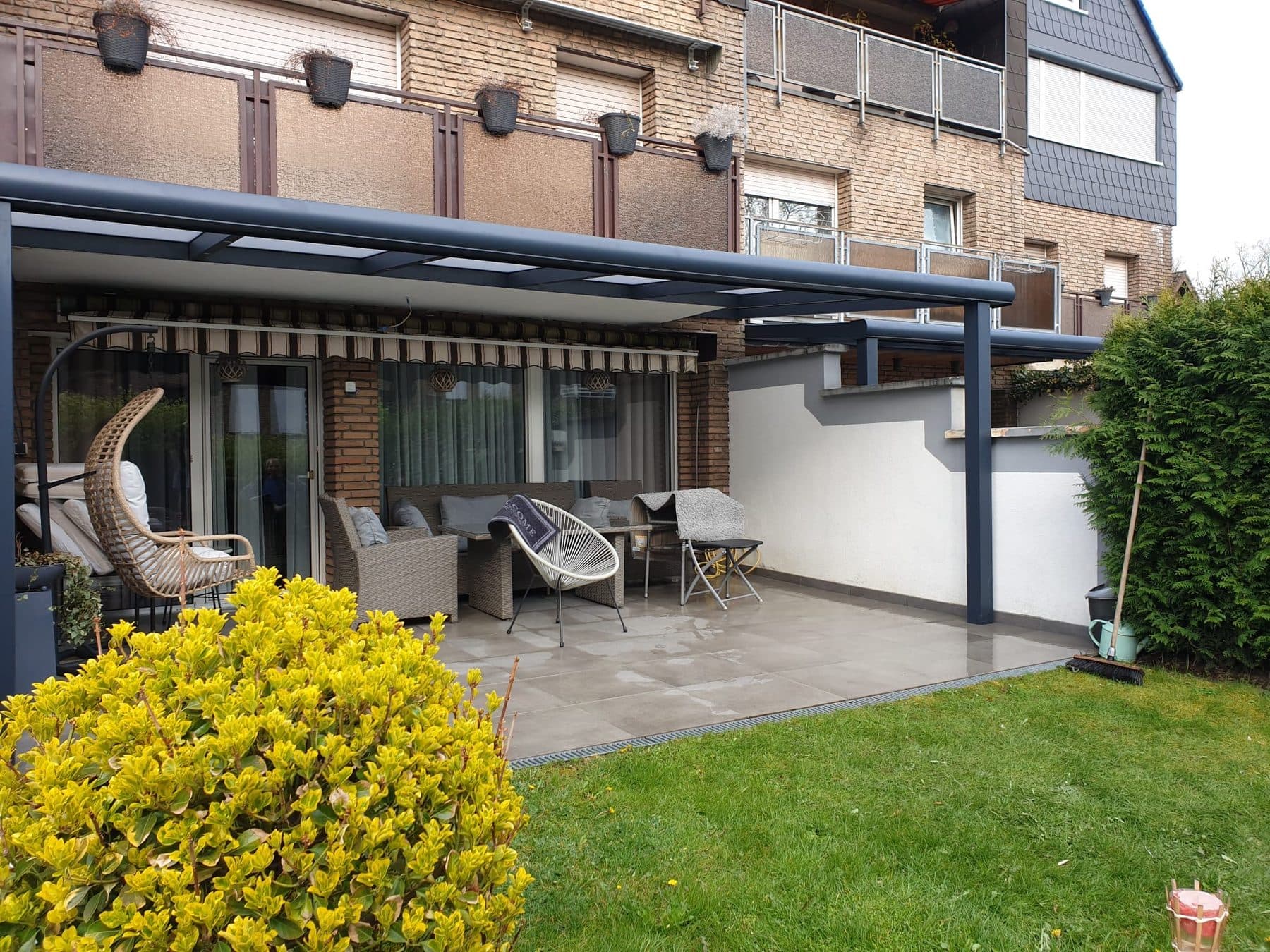 Pergola anthracite à toit polycarbonate opale sur toute la largeur d'une maison en brique, au-dessus d'un salon de jardin et d'un store banne