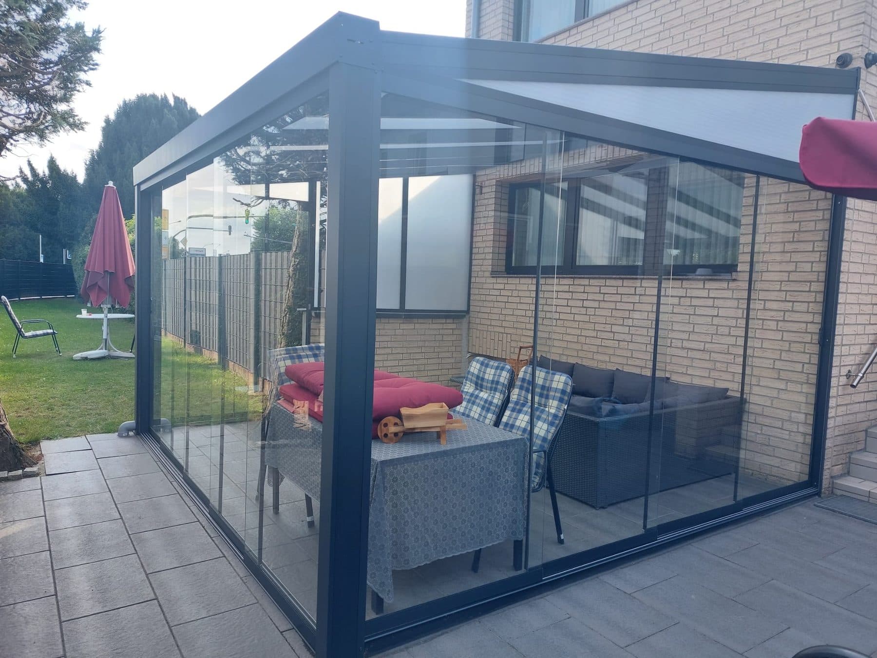 Pergola anthracite fermée par des parois vitrées contre une maison en brique claire, abritant un salon de jardin